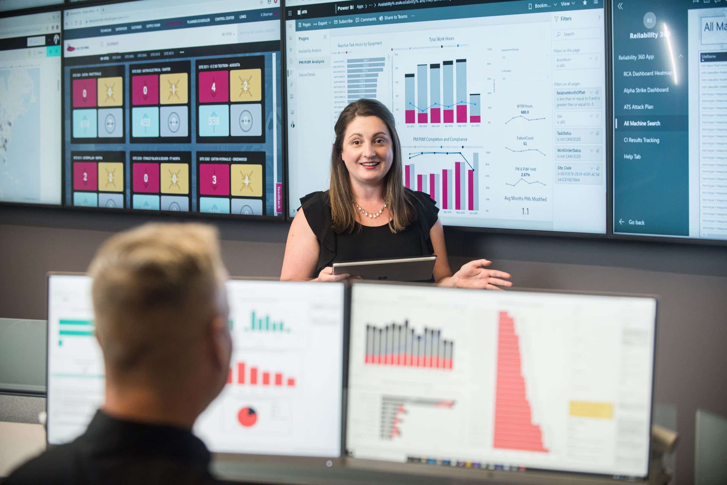 Woman holding tablet in front of screens facing man.