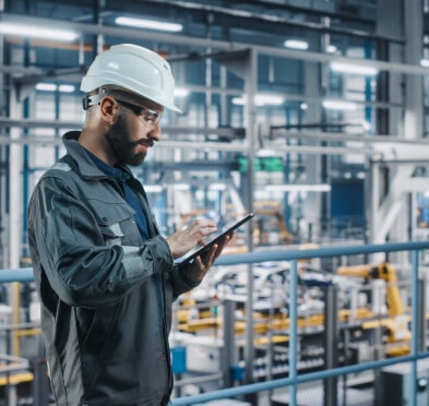 Man wearing blue jumpsuit and white hard hat holding tablet in factory setting