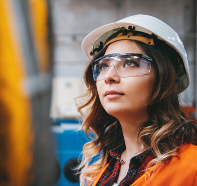 Young woman in orange work vest, safety glasses and hard hat looking at factory equipment