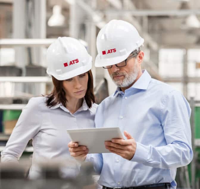 Man and woman in dress clothes and hard hats looking at tablet inside factory