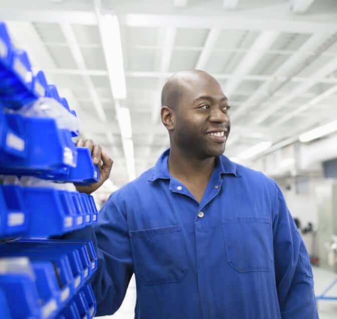 Man in a blue button down looking to the right and touching manufacturing parts
