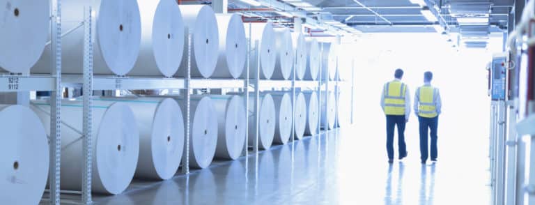 Two men walking through a warehouse holding industrial paper rolls.