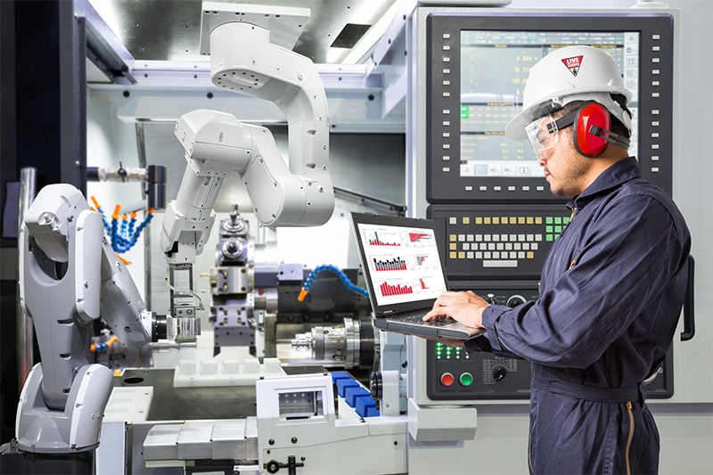 Man in hard hat viewing laptop in front of machinery.