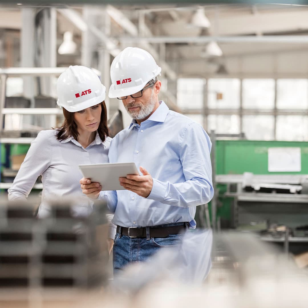 Man and woman in ATS hard hats view tablet in factory