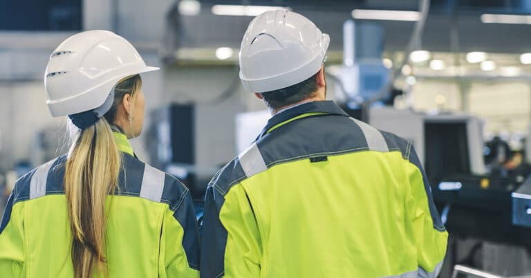 Back of man and woman in factory wearing white hard hats