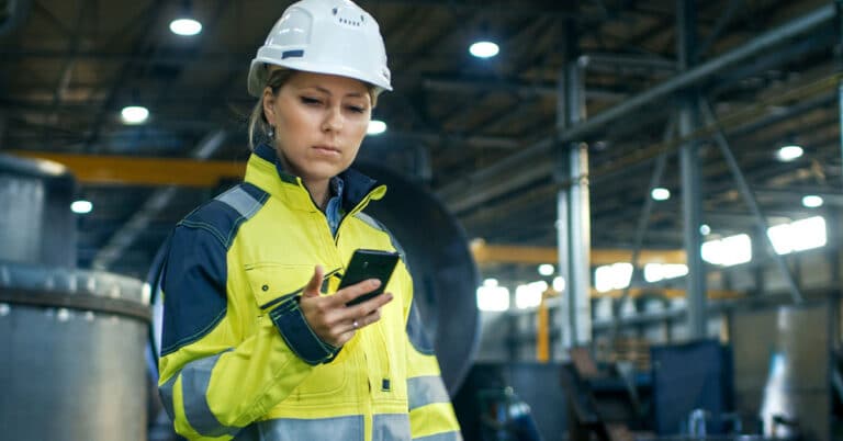 Woman in Factory wearing a white hard Hat looking at mobile Phone.