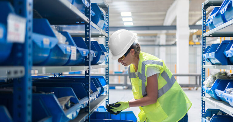 Female warehouse Worker working at a steel Factory wearing a white hard Hat and safety Vest.