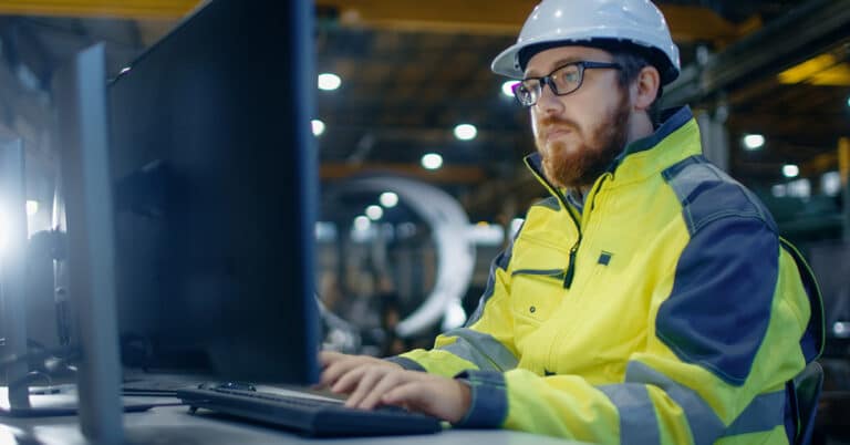 Industrial Engineer Works at Workspace on a Personal Computer. He Wears Hard Hat and Safety Jacket and Works in the Main Workshop of the Heavy Industry Manufacturing Factory.