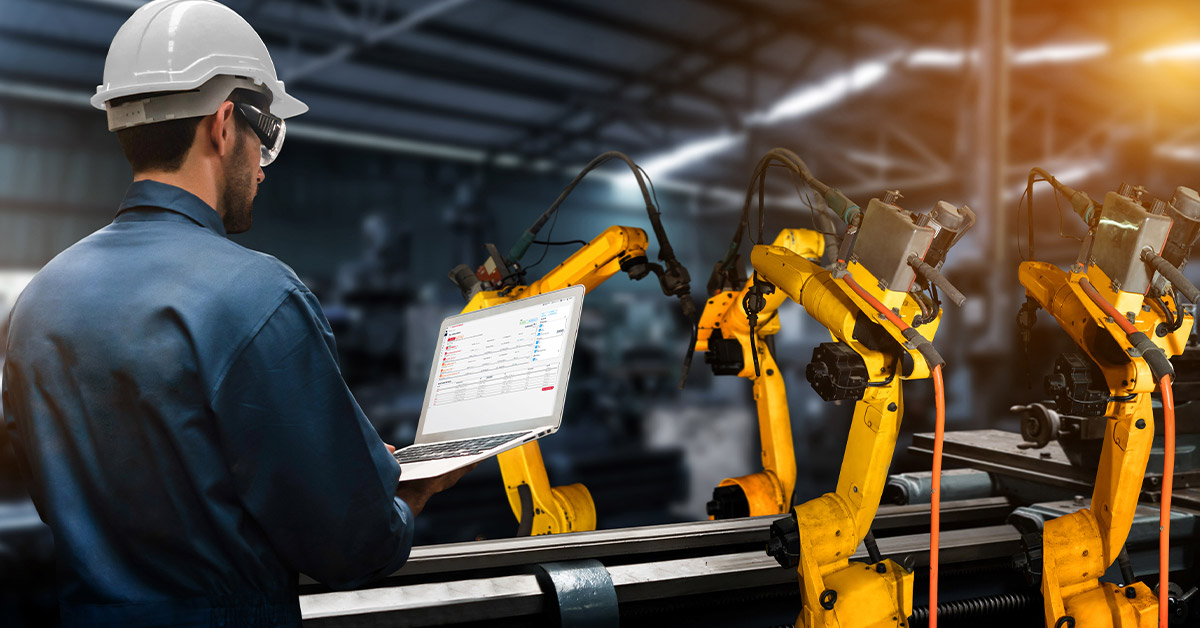 Man standing with laptop next to robotics on the factory floor.