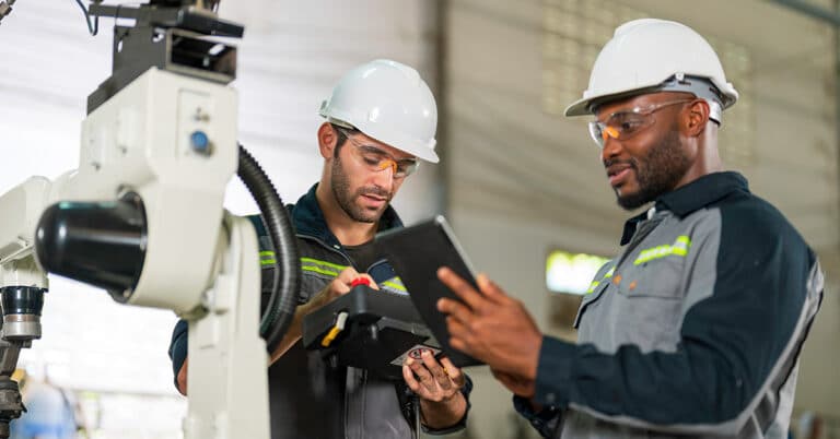 Two industry engineers standing performing maintenance on using robotic controller and tablet in manufacturing facility.
