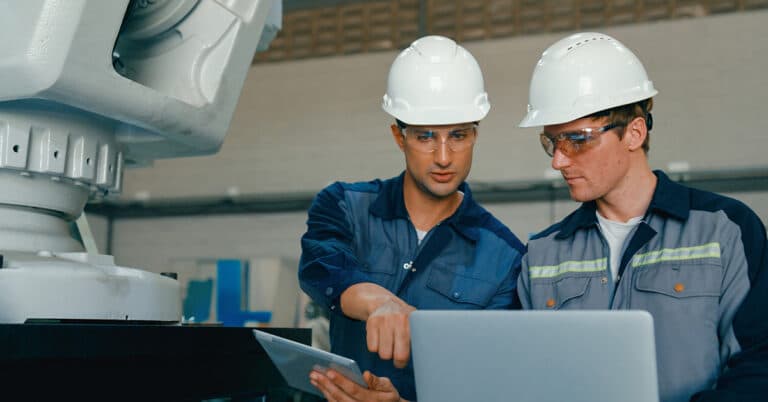 Two young engineers in factory operate large robotic arm, using laptop and tablet to manage the machinery.