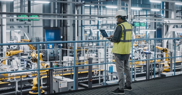 Car factory engineer in high visibility vest using laptop computer in automotive industrial manufacturing facility.