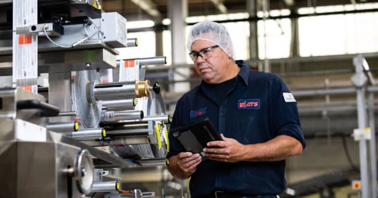 ATS technician with safety glasses and hair net in consumer packaged goods manufacturing facility. Holding tablet.