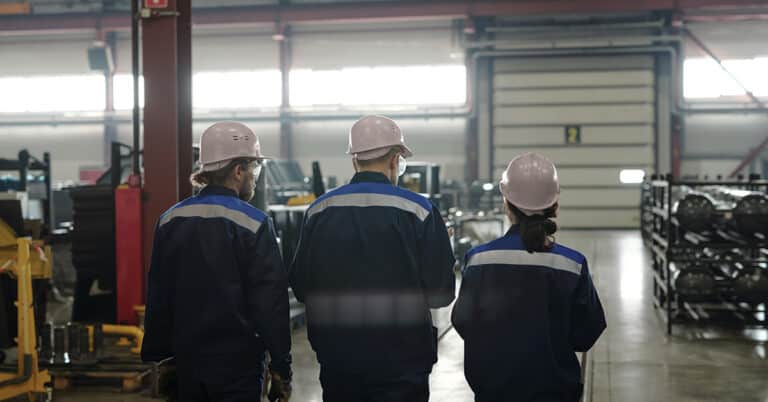 Back view of group of workers or engineers of large contemporary factory moving along workshop with industrial equipment and talking.
