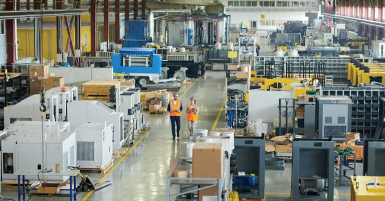 High angle full length portrait of bearded businessman wearing hardhat walking across production workshop accompanied by female factory employee.