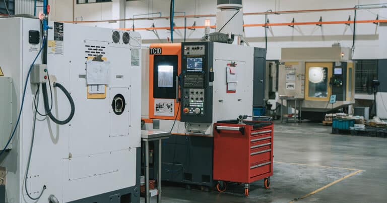 Perspective view down a factory aisle with multiple CNC industrial machines and work tables, illustrating manufacturing production capacity and industrial operations.