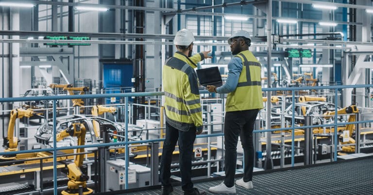 Two Car Factory Specialists in High Visibility Vests Using Laptop Computer.