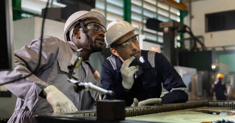 Industrial maintenance technician inspecting machine interior with flashlight in factory. Worker wearing hard hat, safety glasses, gloves performing safety check, troubleshooting and quality control.